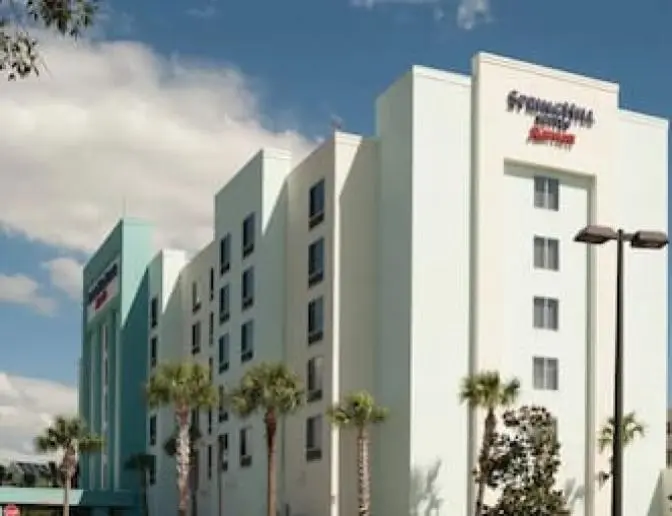 Exterior view of SpringHill Suites by Marriott Orlando International Airport Hotel, a multi-story hotel building with light blue and white walls, surrounded by palm trees and a clear blue sky.