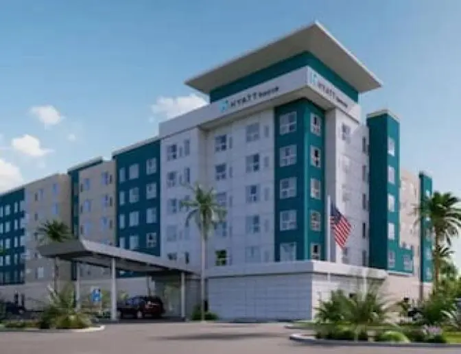 Exterior view of Hyatt House Hotel near Orlando International Airport, featuring a modern multi-story building with teal and white accents, a welcoming entrance, and palm trees under a clear blue sky.