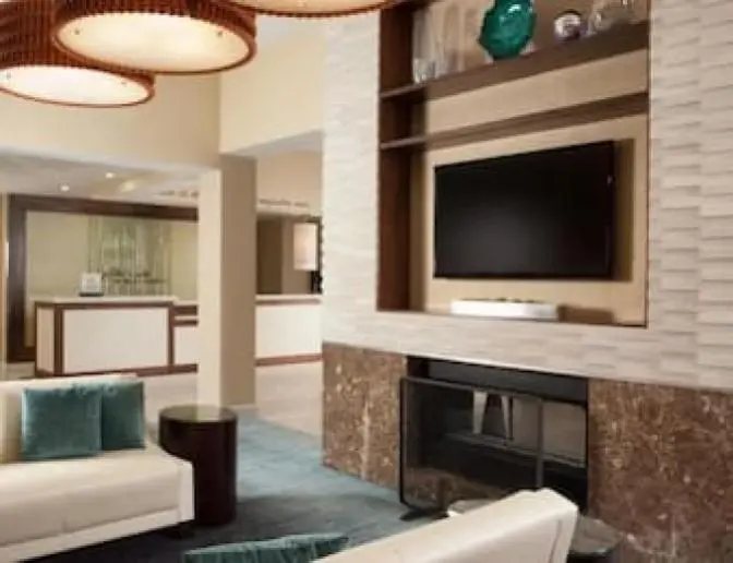 Modern hotel lobby featuring a cozy seating area, flat-screen TV, and fireplace with reception desk in the background.