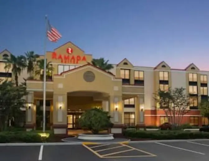 Front view of Ramada Suites Orlando International Airport Hotel at dusk, featuring a beige and red facade with a covered entrance, an American flag, palm trees, and a sunset sky.