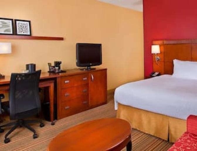 Spacious Courtyard by Marriott hotel room showcasing a king sized bed, a functional work desk with a lamp and chair, a flat-screen TV, and a red armchair with a patterned ottoman.
