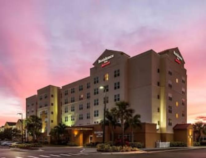 Exterior view of Residence Inn by Marriott Orlando International Airport Hotel at sunset, featuring a multi-story building with illuminated windows and signage, surrounded by palm trees and landscaped grounds.