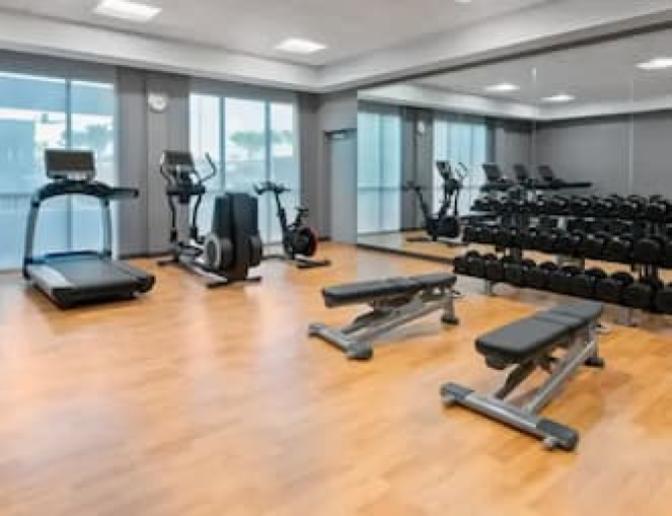 Bright and spacious gym featuring a large open concept gym floor with treadmills, an elliptical and hand weights with benches along a large mirrored wall.