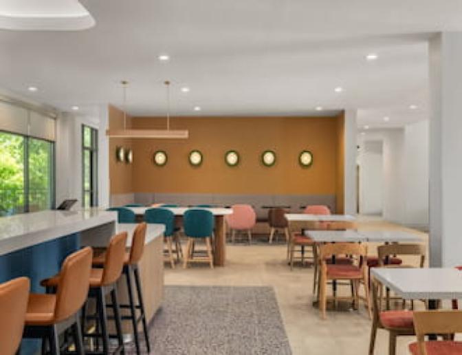 Colorful dining area at EVEN Hotel showcasing a variety of modern chairs, high-top bar seating, and round wall lights against a mustard accent wall, designed for casual social dining.