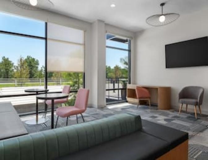 Cozy lounge at EVEN Hotel featuring a sleek black-back sofa, coral accent chairs, a round table, and a flat-screen TV mounted above a compact wooden desk, all framed by tall windows.