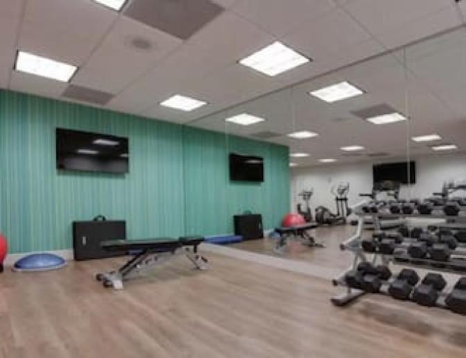 Bright and spacious gym featuring racked hand weights, a bench and medicine balls along a large mirrored wall.