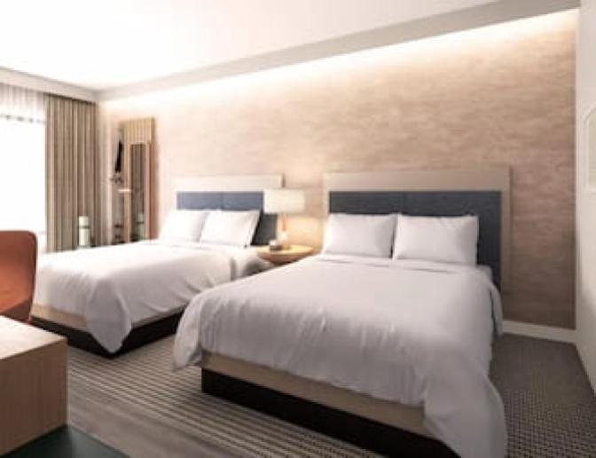 Deluxe EVEN Hotel guest room showcasing two queen beds with upholstered headboards, a textured accent wall, coral desk chair, and wellness wall station with fitness equipment beside a full-length mirror.