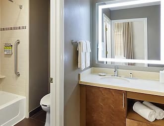 Modern hotel bathroom at Staybridge Suites, including an illuminated mirror, a countertop sink, and a bathtub with shower.
