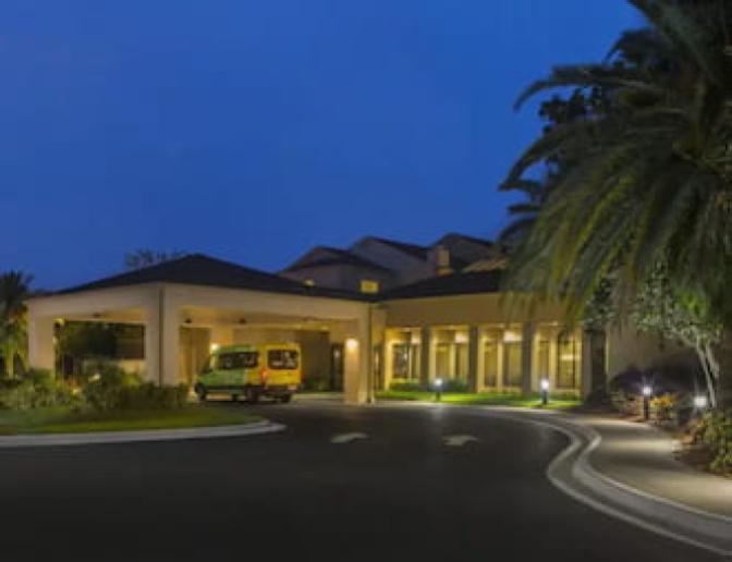 Exterior view of the Courtyard by Marriott Orlando International Airport Hotel at night, featuring a well lit entrance with a yellow shuttle van parked under the covered driveway and surrounded by lush greenery and palm trees
