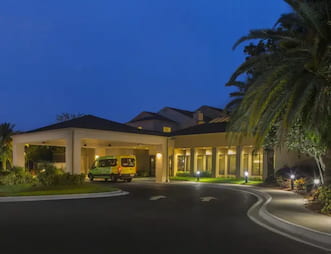 Exterior view of the Courtyard by Marriott Orlando International Airport Hotel at night, featuring a well lit entrance with a yellow shuttle van parked under the covered driveway and surrounded by lush greenery and palm trees
