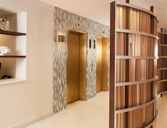 Bright hallway with a white tiled floor, two elevators with gold-colored doors, and a patterned wooden divider.