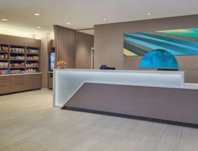Modern hotel lobby with a sleek reception desk, colorful abstract artwork, and a well-stocked snack area featuring shelves of convenience items.
