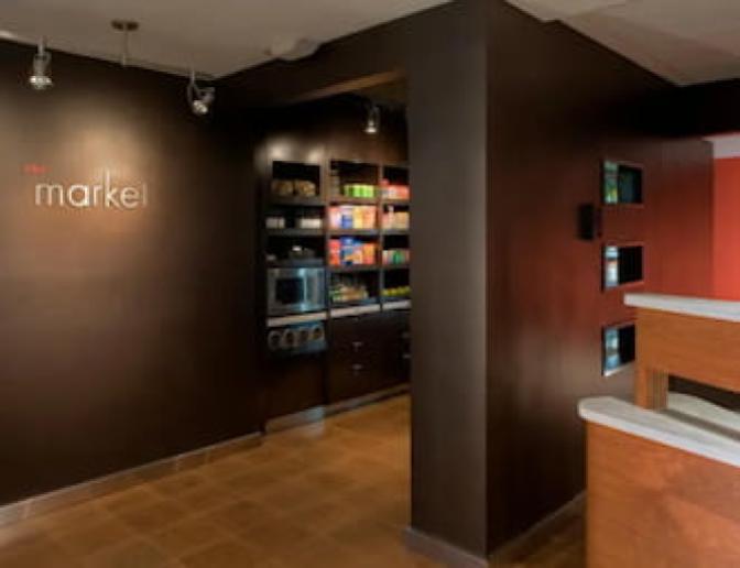 The Market at the Courtyard by Marriott hotel, featuring a selection of snacks, beverages, and convenience items neatly arranged on shelves with a microwave for guest use.