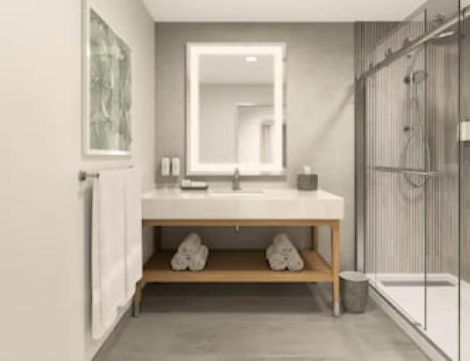 Guest bathroom at EVEN Hotel featuring a sleek vanity with an illuminated mirror, modern sink fixtures, a towel bar with plush white towels, and a glass-enclosed walk-in shower with vertical tile accents.