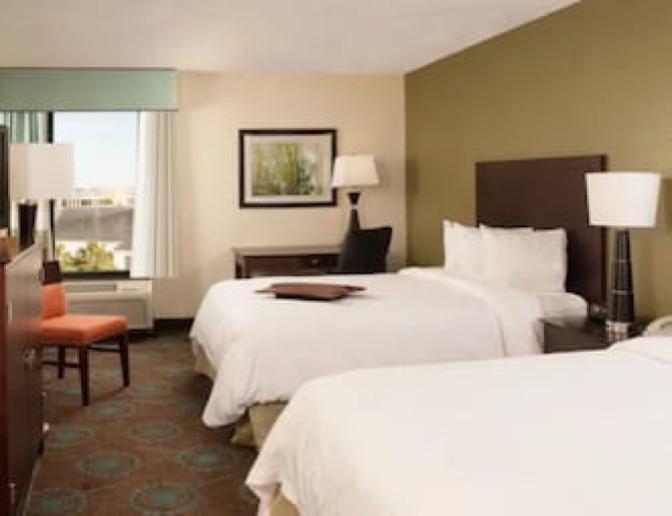 Hotel room with two queen beds, a flat-screen TV, a desk with a chair, and a window with a view. The room features modern decor with dark wood furniture, patterned carpet, and bedside lamps.