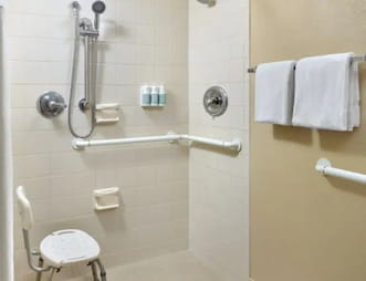 Spacious accessible bathroom in the Courtyard by Marriott hotel, featuring a walk in shower with grab bars, a shower chair, and wall mounted toiletries and towels neatly placed on a towel rack.
