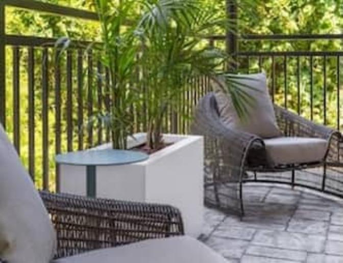 Tranquil outdoor seating area at EVEN Hotel with cushioned wicker chairs, a potted plant on a modern white table, and a private balcony view surrounded by lush green trees.