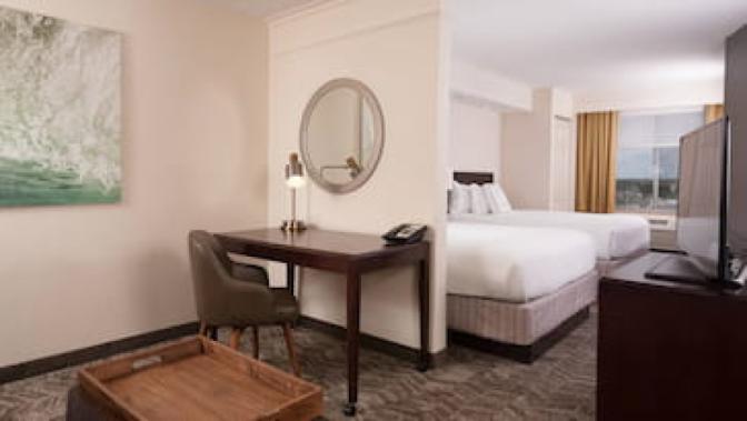 Hotel room at the SpringHill Suites by Marriott featuring two queen beds with white bedding, a desk for work and an ottoman with a wooden tray.