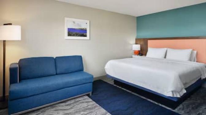 Modern Hampton Inn room featuring  a king-sized bed with a coral and teal accent headboard, a blue upholstered sofa, standing floor lamp, and wall art on a neutral-tone backdrop.