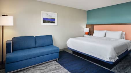 Modern Hampton Inn room featuring  a king-sized bed with a coral and teal accent headboard, a blue upholstered sofa, standing floor lamp, and wall art on a neutral-tone backdrop.