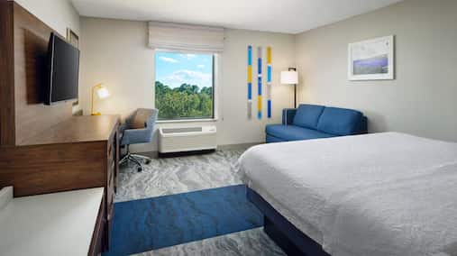 Hotel room at Hampton Inn with a king-sized bed, blue upholstered sofa, work desk with ergonomic chair and gold task lamp, flat-screen TV, and a large window with views of treetops and blue sky.