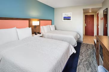 Hampton Inn hotel room featuring two queen beds with coral and teal accent headboards, crisp white linens, a nightstand with a lamp, flat-screen TV, and wood-tone furnishings with a view toward the entry hallway.