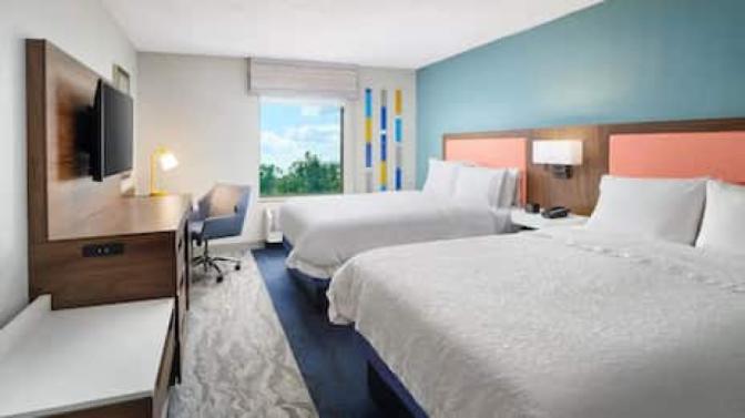 Hampton Inn guest room featuring two queen beds with coral accent headboards, a work desk with ergonomic chair and task lamp, flat-screen TV, and a large window with scenic views of trees and sky.