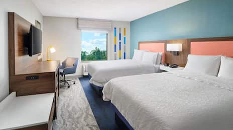 Hampton Inn guest room featuring two queen beds with coral accent headboards, a work desk with ergonomic chair and task lamp, flat-screen TV, and a large window with scenic views of trees and sky.