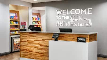 Hampton Inn lobby reception area featuring a wood-paneled front desk with dual monitors, a snack shop stocked with beverages and treats, and a wall sign that reads “Welcome to the Sunshine State” with a Florida state outline.