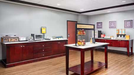 Hampton Inn breakfast area featuring wood cabinetry, a central island with pastry displays, a toaster station, waffle maker, microwave, juice dispensers, and coffee setup, all set against neutral walls and wood-style flooring.