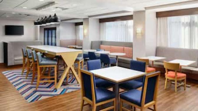 Hampton Inn breakfast seating area featuring a mix of dining tables, high-top communal seating with pendant lighting, upholstered booths, and colorful chairs set on wood-style flooring with a vibrant patterned rug.