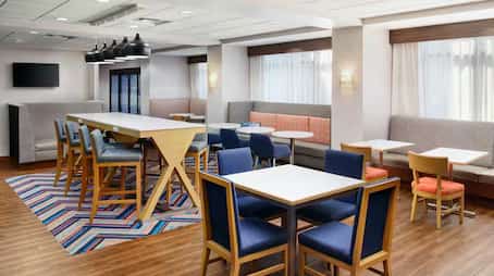Hampton Inn breakfast seating area featuring a mix of dining tables, high-top communal seating with pendant lighting, upholstered booths, and colorful chairs set on wood-style flooring with a vibrant patterned rug.
