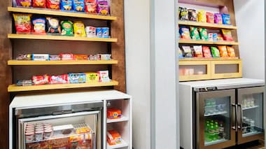 Hampton Inn suite shop featuring open shelves stocked with snacks, chips, and candy, along with refrigerated coolers displaying bottled beverages, yogurt, and ready-to-eat items.