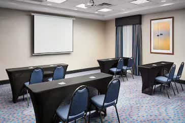 Hampton Inn meeting room featuring several rectangular tables draped in black linens, blue cushioned chairs, a ceiling-mounted projector with a dropdown screen, and a large window with patterned curtains.