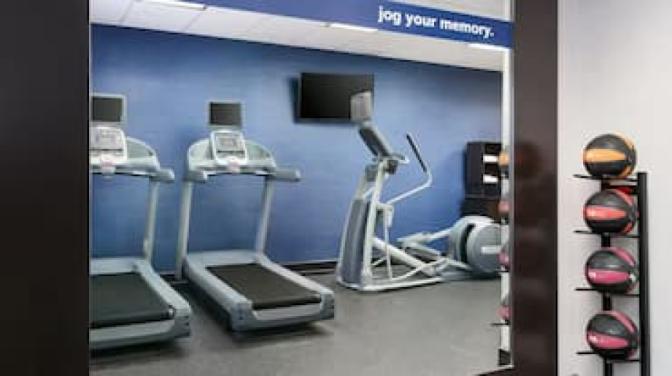 Hampton Inn fitness center featuring two treadmills, an elliptical machine, a flat-screen TV, a set of medicine balls on a vertical rack, and a blue accent wall with the phrase "jog your memory."