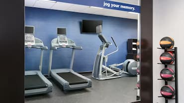 Hampton Inn fitness center featuring two treadmills, an elliptical machine, a flat-screen TV, a set of medicine balls on a vertical rack, and a blue accent wall with the phrase "jog your memory."