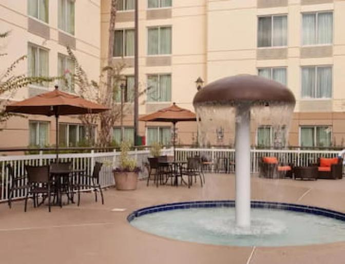 Courtyard with a splash pad water fountain, tables and chairs under umbrellas, and seating areas with orange cushions.