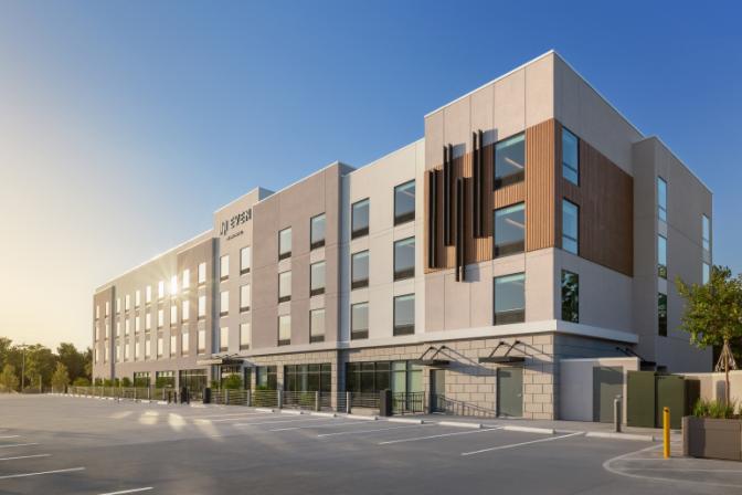 Modern exterior of EVEN Hotel during daylight, showcasing a four-story building with geometric design elements, large grid windows, and an expansive guest parking area.