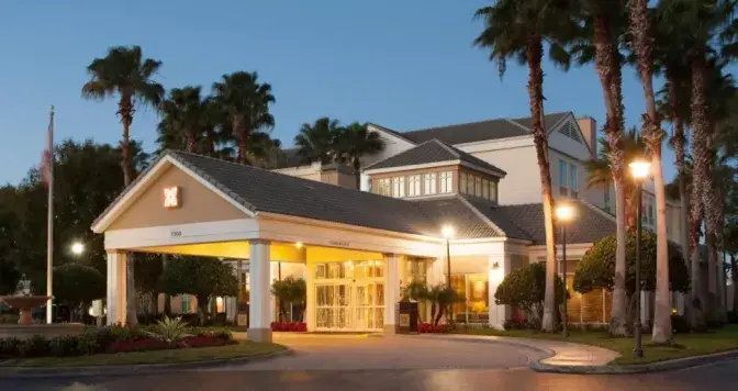 Exterior view of Hilton Garden Inn Orlando Airport Hotel entrance at dusk, surrounded by tall palm trees and illuminated by warm lighting.