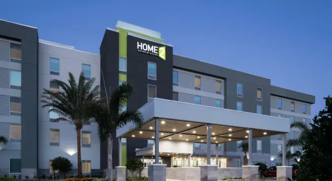 Exterior view of Home 2 Suites Orlando Airport Hotel, featuring a large modern grey and white multistory building with lush greenery and tall palm trees under an evening sky.