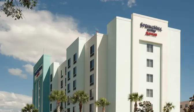 Exterior view of SpringHill Suites by Marriott Orlando International Airport Hotel, a multi-story hotel building with light blue and white walls, surrounded by palm trees and a clear blue sky.