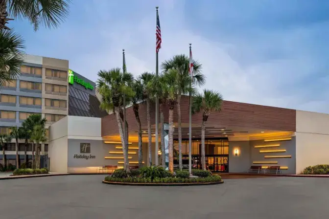 Exterior view of Holiday Inn Orlando International Airport, featuring a modern entrance. with warm lighting, tall palm trees, and flags displayed against an evening sky.