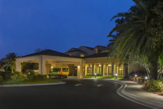 Exterior view of the Courtyard by Marriott Orlando International Airport Hotel at night, featuring a well lit entrance with a yellow shuttle van parked under the covered driveway and surrounded by lush greenery and palm trees