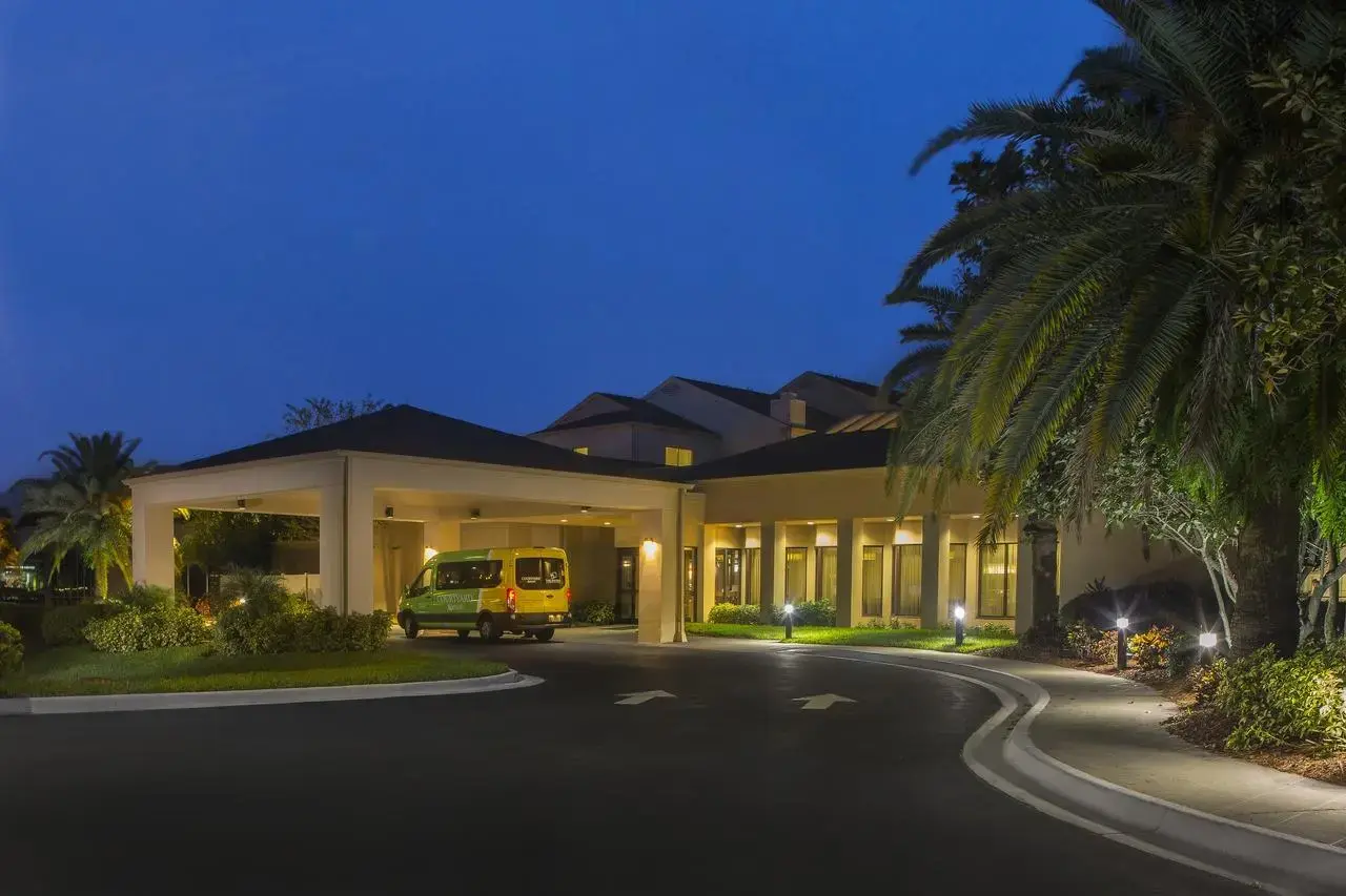 Exterior view of the Courtyard by Marriott Orlando International Airport Hotel at night, featuring a well lit entrance with a yellow shuttle van parked under the covered driveway and surrounded by lush greenery and palm trees