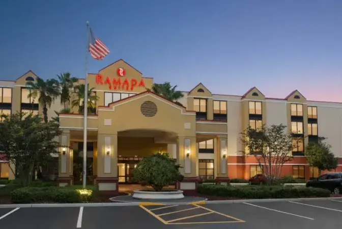 Front view of Ramada Suites Orlando International Airport Hotel at dusk, featuring a beige and red facade with a covered entrance, an American flag, palm trees, and a sunset sky.