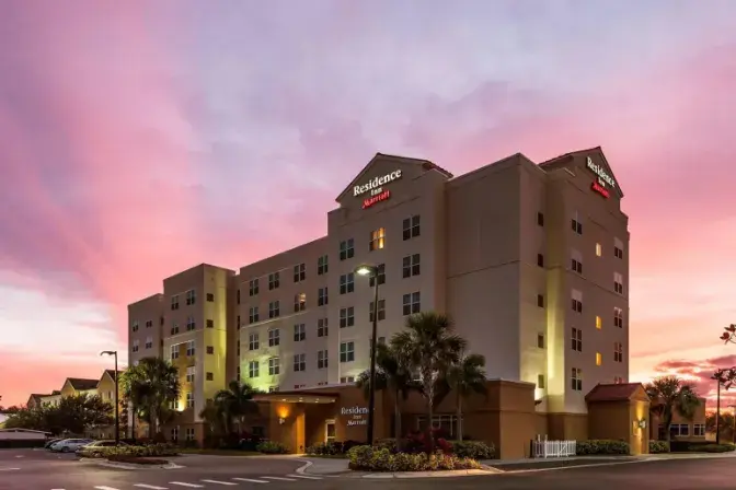 Exterior view of Residence Inn by Marriott Orlando International Airport Hotel at sunset, featuring a multi-story building with illuminated windows and signage, surrounded by palm trees and landscaped grounds.