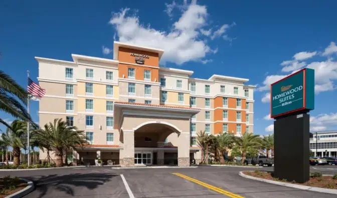 Exterior view of Homewood Suites by Hilton featuring a multi-story building with a beige and orange facade, a covered entrance, and surrounding palm trees.