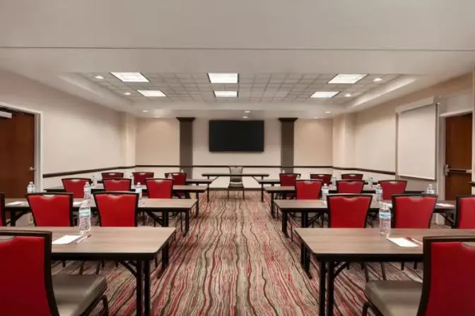 Spacious business center at Ramada Suites featuring a large television at the front of the room with many tables and chairs set up.
