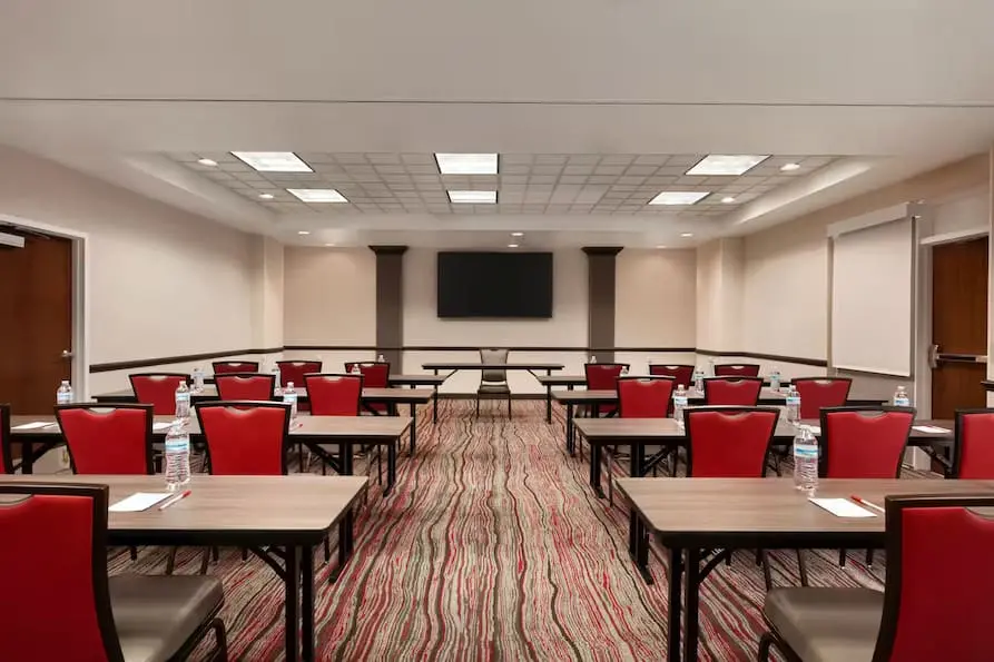 Spacious business center at Ramada Suites featuring a large television at the front of the room with many tables and chairs set up.