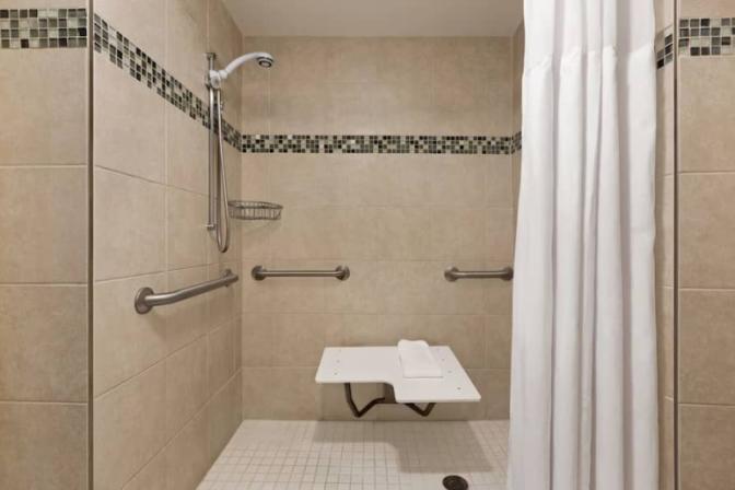 Bathroom inside a suite at the Ramada Suites Orlando International Airport Hotel, featuring an accessible shower with bench.